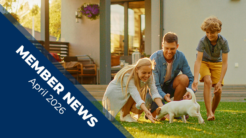 Four people kneel and crouch on a sunlit lawn, reaching toward a small white puppy, with a modern house, deck, outdoor furniture, and trees in the background.