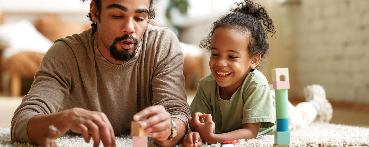 An adult and a young child lie on a carpeted floor indoors, placing wooden blocks together to build a small structure. Additional toy blocks are visible nearby.