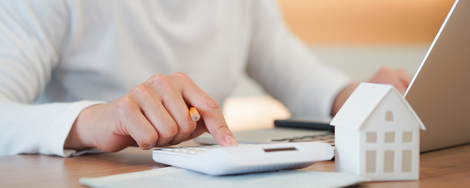 A close-up of hands using a calculator on a desk beside a small white model house, with a laptop partially visible in the background.