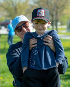 An adult stands outdoors holding a small child in front of them. The child wears a dark long‑sleeve shirt with a recycling graphic and a black cap featuring a cartoon character patch. Trees and grass are visible in the background.