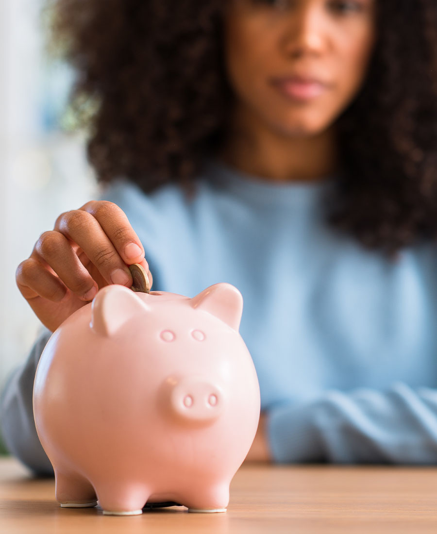 A close-up view of a person placing a coin into a pink piggy bank while holding the piggy bank steady with one hand. The background is softly out of focus.