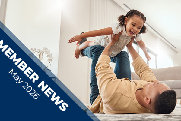 An adult lies on a carpeted living room floor lifting a small child horizontally above the adult’s chest with both hands. A diagonal blue banner on the left reads “Member News, May 2026.”