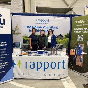 Three adults stand behind a display table at an indoor event booth. Banners behind them read “rapport, a division of WFCU” and “The Home You Want is Within Reach.” The table covering reads “rapport credit union,” with promotional items displayed.