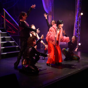 A stage performance shows one performer standing at a microphone wearing a bright outfit, while several other performers in dark costumes kneel or stand around them with arms raised. Stage lighting and stairs are visible in the background.