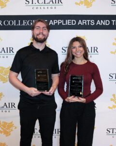 Two adults stand holding glass award plaques in front of a banner backdrop. The backdrop reads “St. Clair College” and “Centre for the Arts and Technology.” The plaques display WFCU Credit Union and St. Clair College branding.