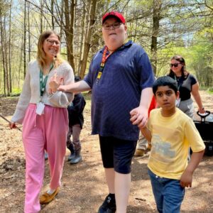 A group walks along a wooded trail outdoors. In the foreground, one adult walks between two children while holding hands. Other adults walk behind them. Trees, fallen leaves, and a dirt path are visible.