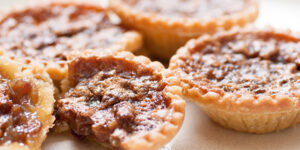 Close-up photograph of several butter tarts with golden pastry shells and glossy brown filling, arranged closely together.