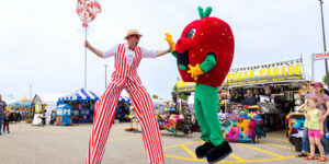 Outdoor festival scene showing a performer on stilts interacting with a large strawberry mascot. Colourful vendor booths and fair attractions line the background, with attendees watching nearby.