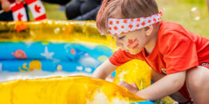 A young child playing with water in a shallow inflatable pool outdoors, wearing a red shirt and a headband with red maple leaves. The scene suggests a summer activity.