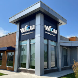 Exterior view of a modern WFCU Banking Centre with blue and grey exterior panels, large windows, and WFCU signage displayed on two sides of the corner façade under a clear sky.