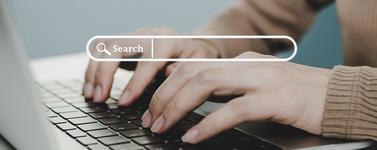 Close-up of hands typing on a laptop keyboard with a search bar graphic displayed above. The search bar contains the word 'Search' and represents an online search or information lookup process.