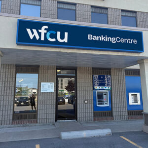 Exterior view of a WFCU Banking Centre Kingston branch showing the main entrance, windows, and automated banking machine. A blue sign above the entrance displays the text 'WFCU BankingCentre'.