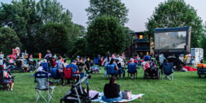 Community members seated on lawn chairs and blankets in a park watching a movie during City of London movie night event on a large outdoor screen at dusk.