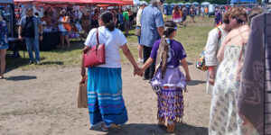 Two attendees walk hand in hand through the Caldwell First Nation: 2nd Annual Competition Pow Wow in Essex, with vendor booths and event participants visible in the background.