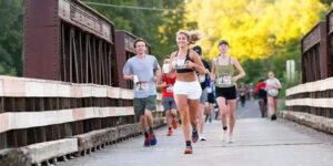 Participants in a community road race run across a pedestrian bridge during Royal Canadian Legion: Kakabeka Falls Half Marathon and 8K.