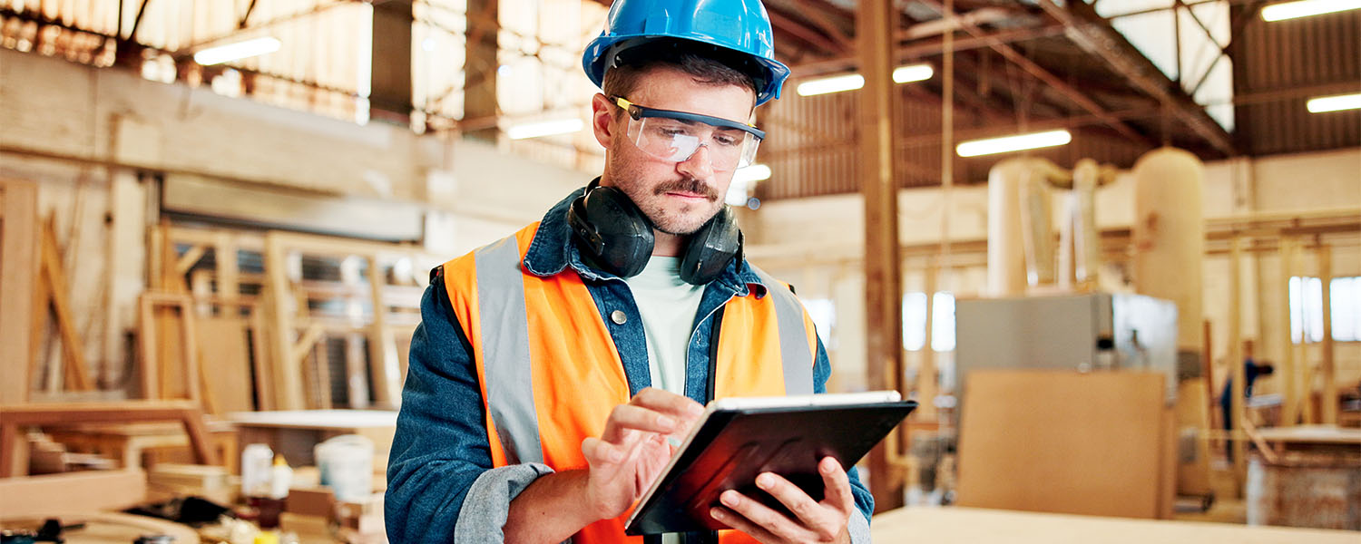 Person wearing safety equipment, including a hard hat, safety glasses, and hearing protection, using a tablet in a manufacturing facility. Industrial equipment and workshop materials are visible in the background.