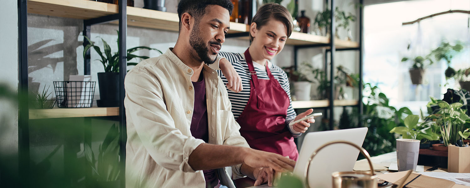 Person using a laptop while another person stands nearby holding a smartphone in a plant-filled small business workspace. Shelving with plants and supplies is visible in the background.