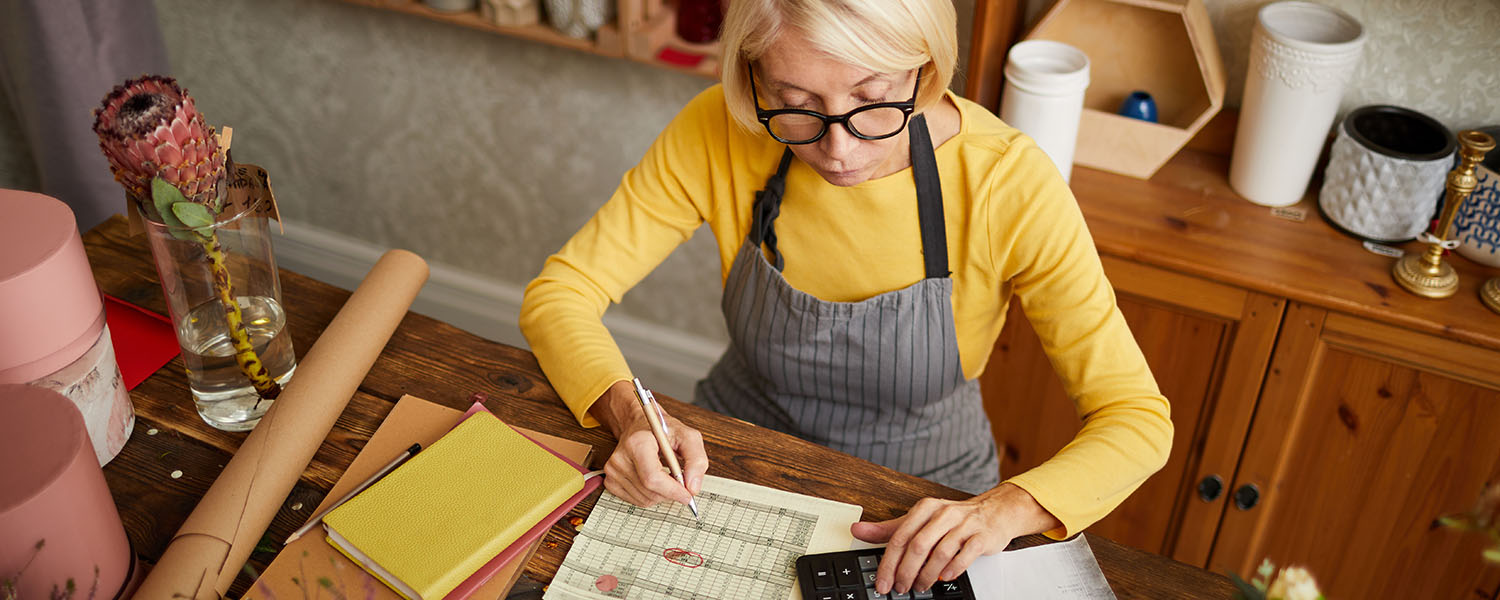 Person wearing an apron and glasses reviewing paperwork and using a calculator at a wooden table in a home-based business workspace. Notebooks, documents, and packaging materials are visible on the table.